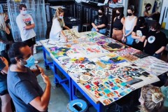 一群大学生的中心是一张摆满彩色艺术品的桌子，他们看着一位举手说话的老师。所有人都戴上口罩。