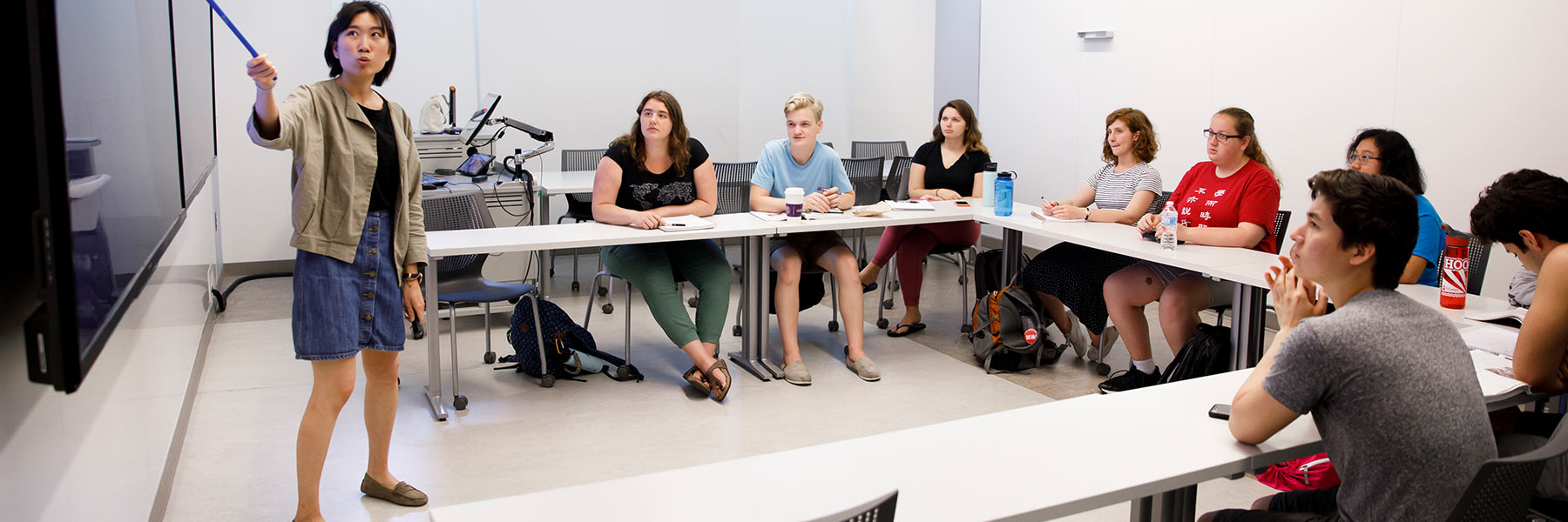 一名教师指着墙上的屏幕，学生们坐在教室里听着看着。